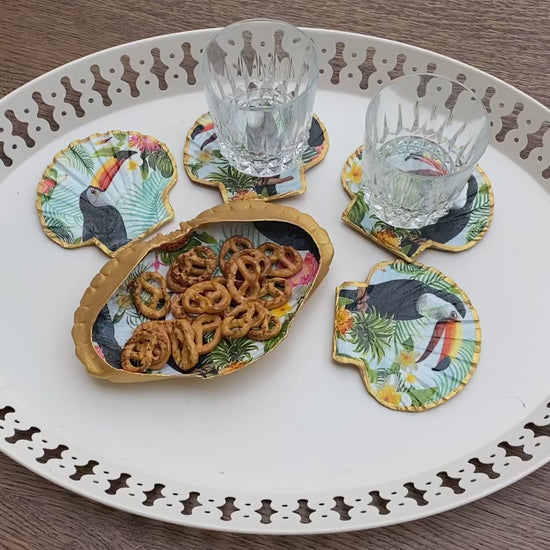 Des sous-verres uniques en coquilles Saint-Jacques, décorés à la main avec un liseré doré et un motif toucan vibrant, apportant une touche tropicale et élégante à votre table.